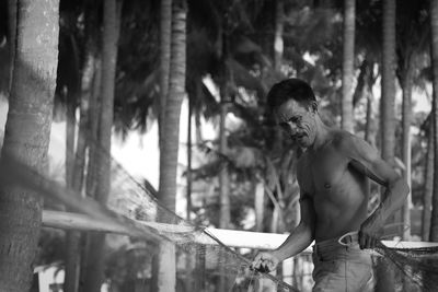 Shirtless man holding fishing net while standing against trees