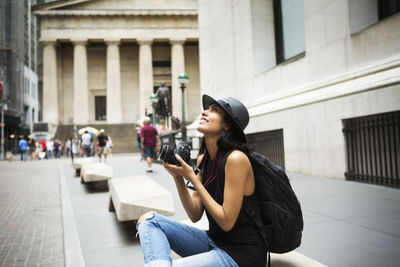 Woman with camera carrying backpack sitting on bench in city