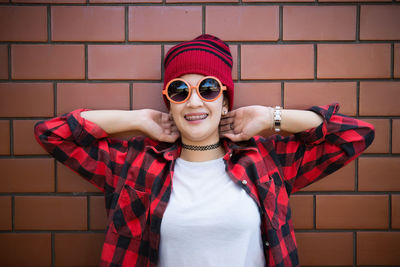 Portrait of smiling woman in sunglasses and knit hat standing against brick wall