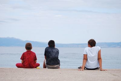 Rear view of boys sitting by sea against sky