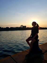 Woman sitting by lake against sky during sunset