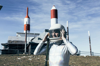 Man in spacesuit standing on rocky ground against metal fence and striped rocket shaped antennas on sunny day