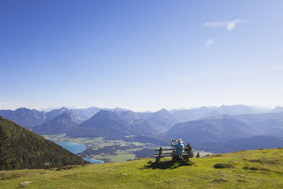 Rear view of senior male hiker photographing while sitting on bench while looking at dachstein mountains against blue sky