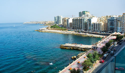 Sliema rocky coastline, malta, with cafes, modern buildings, and bridge at the distance.