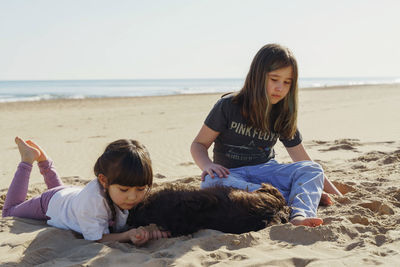 Side view of two girls sitting at beach and playing with dog