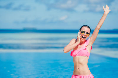 Portrait of young woman with arms raised standing at beach against sky