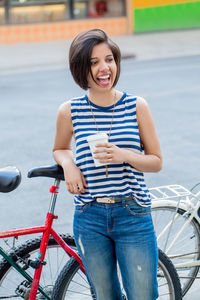Cheerful young woman drinking coffee while standing by bicycle