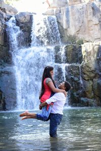 Full length of young man lifting woman while standing against waterfall at river