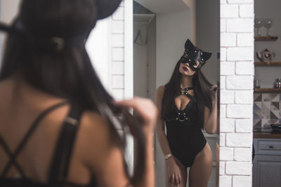 Rear view of sensuous woman wearing mask standing in front of mirror at home