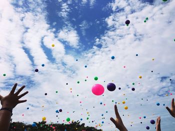 Cropped image of hands reaching towards colorful balloons flying against cloudy sky