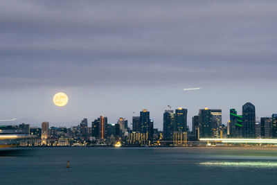 Sea by illuminated modern city against full moon in sky at dusk