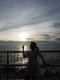 Rear view of man standing by railing against sea during sunset