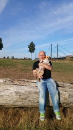 Full length of man carrying dog while sitting on log against sky