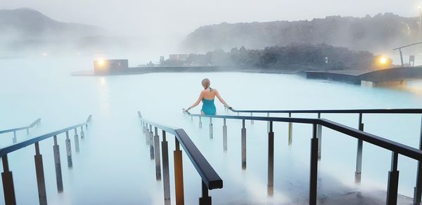 Rear view of woman sitting on railing amidst smoke by lake