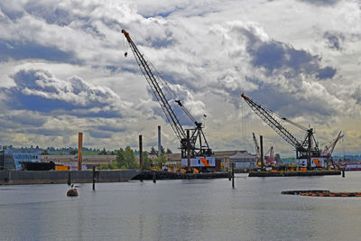 Cranes at commercial dock against cloudy sky