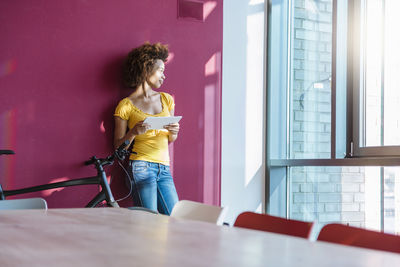 Young woman standing in front of read wall, looking out of window, holding digital tablet