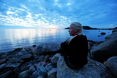 Woman sitting on rock at sea shore against sky at dusk