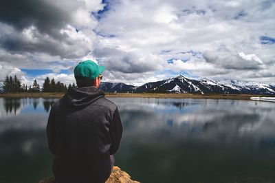 Rear view of man sitting on rock by lake against cloudy sky