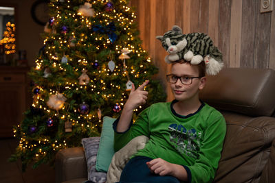 Portrait of boy with toy animal on head sitting against christmas tree at home