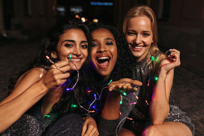 Portrait of smiling friends with illuminated lighting equipment sitting in city at night