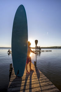 Man with paddleboard standing at pier during sunset