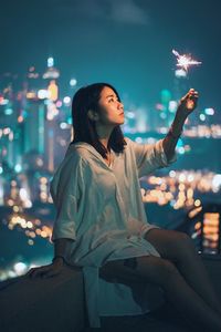 Young woman holding sparkler in city against sky at night