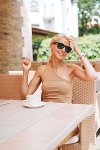 Adult elegant blonde woman in sunglasses beige clothes with cup of coffee in summer city street cafe