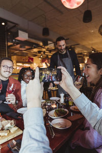 Cropped image of man taking picture of male and female friends through mobile phone in bar