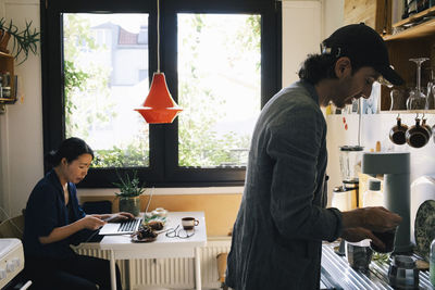 Male architect preparing coffee while female colleague using smart phone in home office