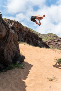 Shirtless male touching bare feet while leaping from cliff on sandy beach against cloudy blue sky on sunny summer day