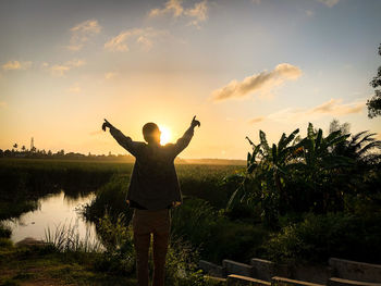 Rear view of man with arms raised standing against sky during sunset