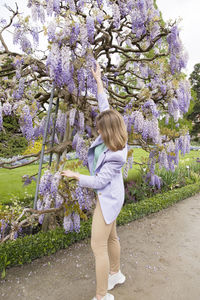 A young woman in a lilac jacket poses near the flowering wisteria in the garden