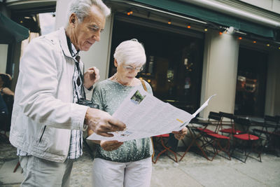 Senior couple reading map while standing on sidewalk in city during vacation