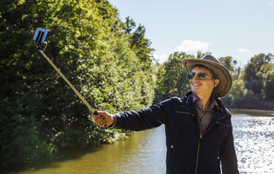 Young man wearing sunglasses taking selfie while standing by lake