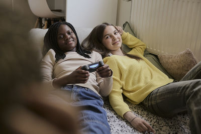 Smiling teenage girl holding game controller while lying down with female friend in bedroom