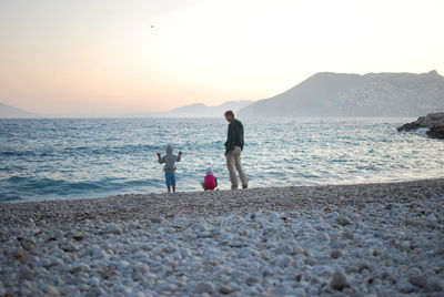 Rear view of father with two kids walking at beach against sky during sunset