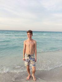 Portrait of shirtless man standing at beach against sky