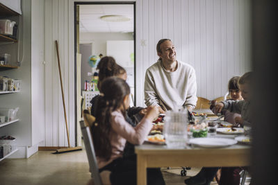 Happy male child care worker with students having breakfast at table in kindergarten