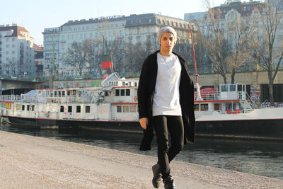 Portrait of young man standing on promenade by boat moored at river in city