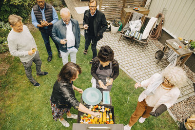 High angle view of smiling senior men looking at females preparing dinner on barbecue during garden party