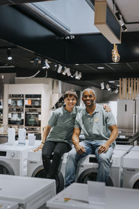 Portrait of smiling male and female retail clerks sitting on washing machines in electronics store