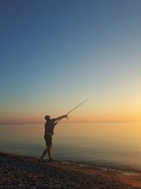 Side view of man fishing in sea against sky during sunset