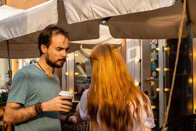 Young couple talking while standing near street cafe and drinking takeaway coffee