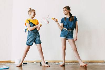 A girl with her sister in bright blue and yellow clothes helps to paint the walls in her room white.
