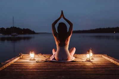 Rear view of woman practicing yoga while sitting on pier by sea against sky