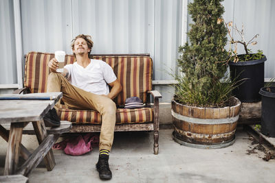 Relaxed carpenter holding disposable cup while sitting on sofa outside workshop