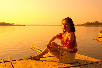 Woman sitting on wooden raft in lake against clear sky during sunset