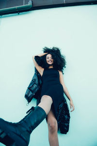 From below of smiling young ethnic female with afro hair wearing trendy black dress and jacket leaning on stone building wall while standing in city street in daytime