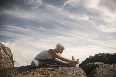 Blond flexible woman practicing yoga on rock formation at beach against sky