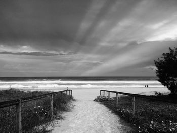 Walkway leading towards beach against cloudy sky during sunset